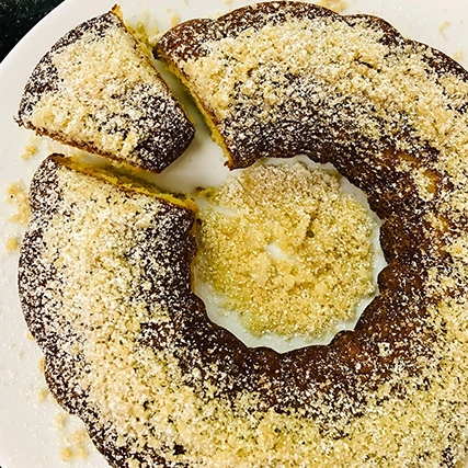 A round cake with a slice cut out, topped with powdered sugar, on a white plate. The cake has a golden-brown crust and a soft, moist interior.