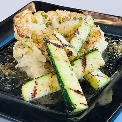 Grilled zucchini slices on a roasted cauliflower steak, garnished with herbs and served on a black square plate with creamy sauce.