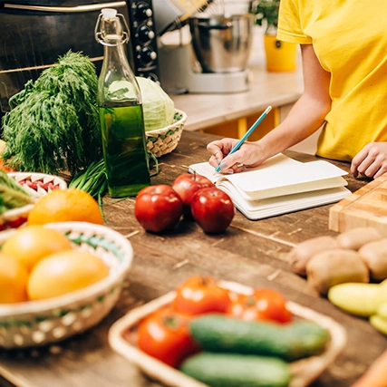 Person writes in a notebook at a kitchen counter with vegetables, fruits, and a bottle of oil.