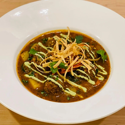 A bowl of soup or stew with meatballs, vegetables, a drizzle of green sauce, and crispy tortilla strips, presented in an appealing mix of colors and textures."