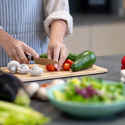 A person slicing cherry tomatoes on a wooden cutting board with other vegetables around.