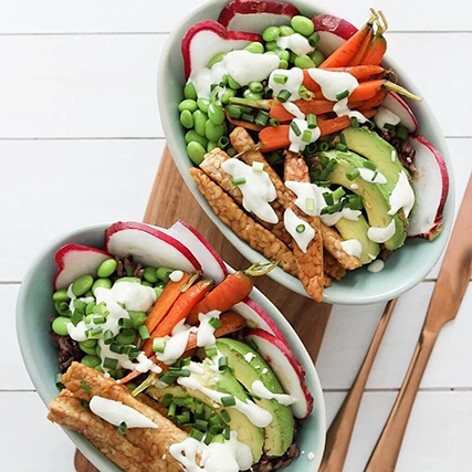 Two light blue bowls filled with a colorful salad including tempeh, avocado slices, baby carrots, edamame, radish slices, and a creamy white dressing, arranged on a wooden board.