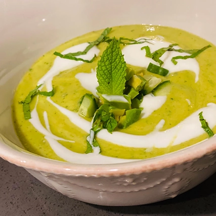 Close-up of a bowl of creamy green soup garnished with swirls of white cream, chopped herbs, and a sprig of mint.