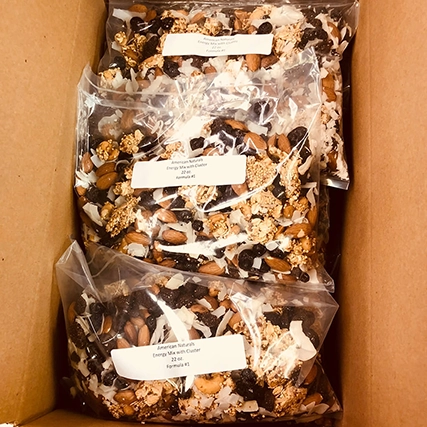 A mix of nuts, seeds, dried fruits, and granola clusters, packaged in three clear plastic bags with white labels, inside a cardboard box.
