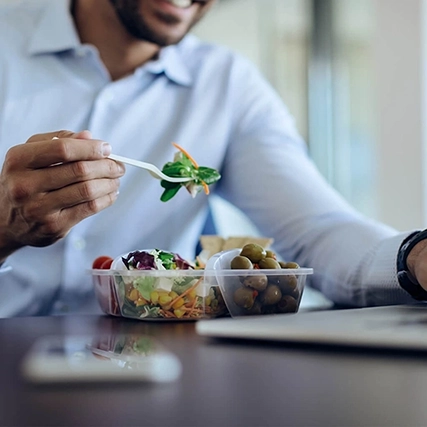A person eating a salad at a desk while working on a laptop, with a plastic container holding the salad.