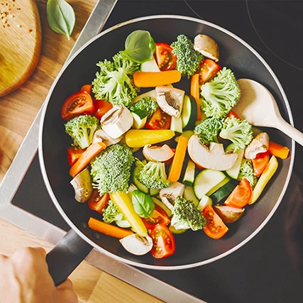 Colorful assortment of vegetables, including broccoli, mushrooms, cherry tomatoes, carrots, and zucchini, being cooked in a frying pan on a stovetop with a wooden spoon.